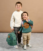 A boy holding the Lassig mini backpack for kids in a dark green colour, with his arm around a younger boy who's wearing the lilac version.
