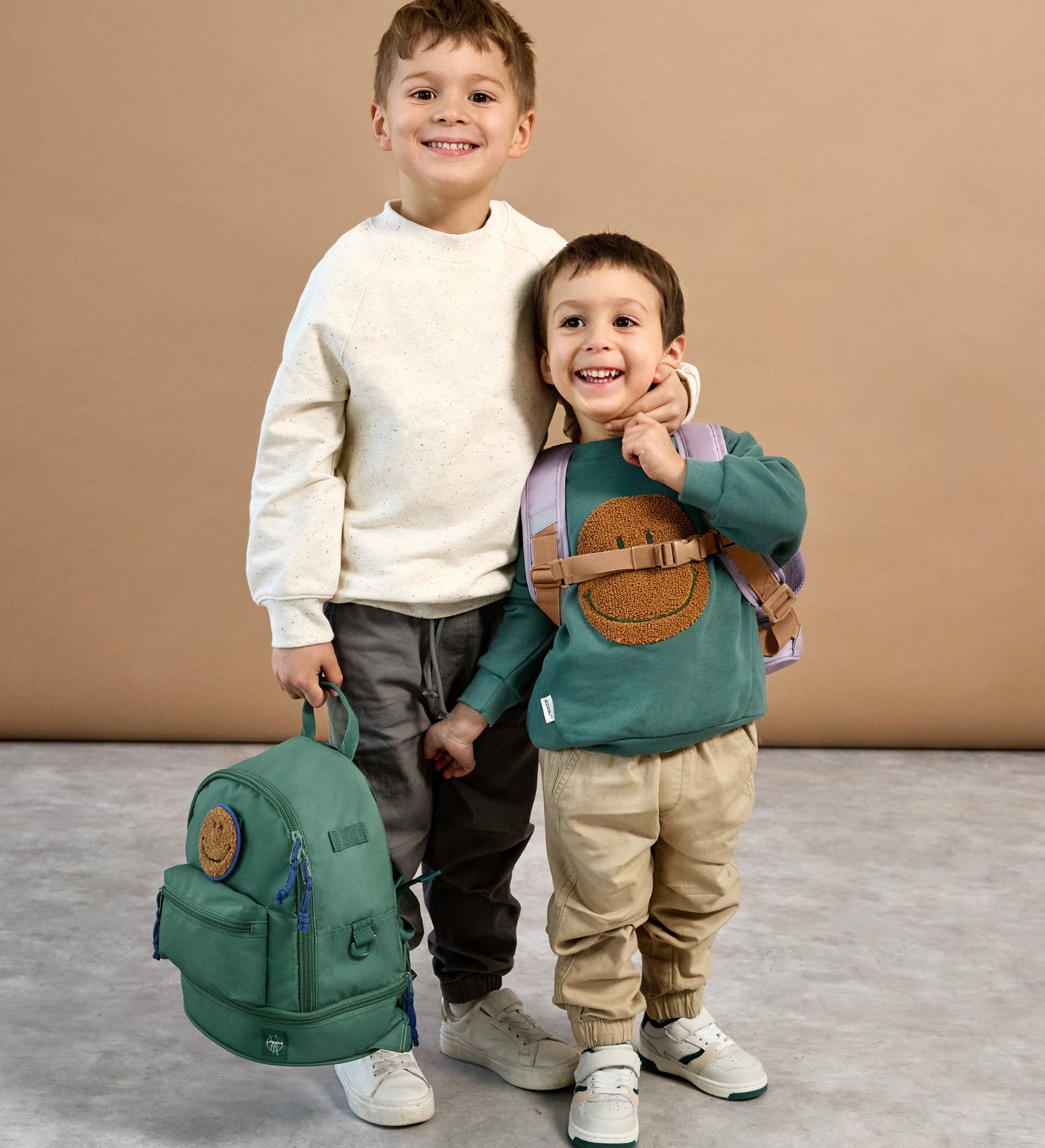 A boy holding the Lassig mini backpack for kids in a dark green colour, with his arm around a younger boy who's wearing the lilac version.
