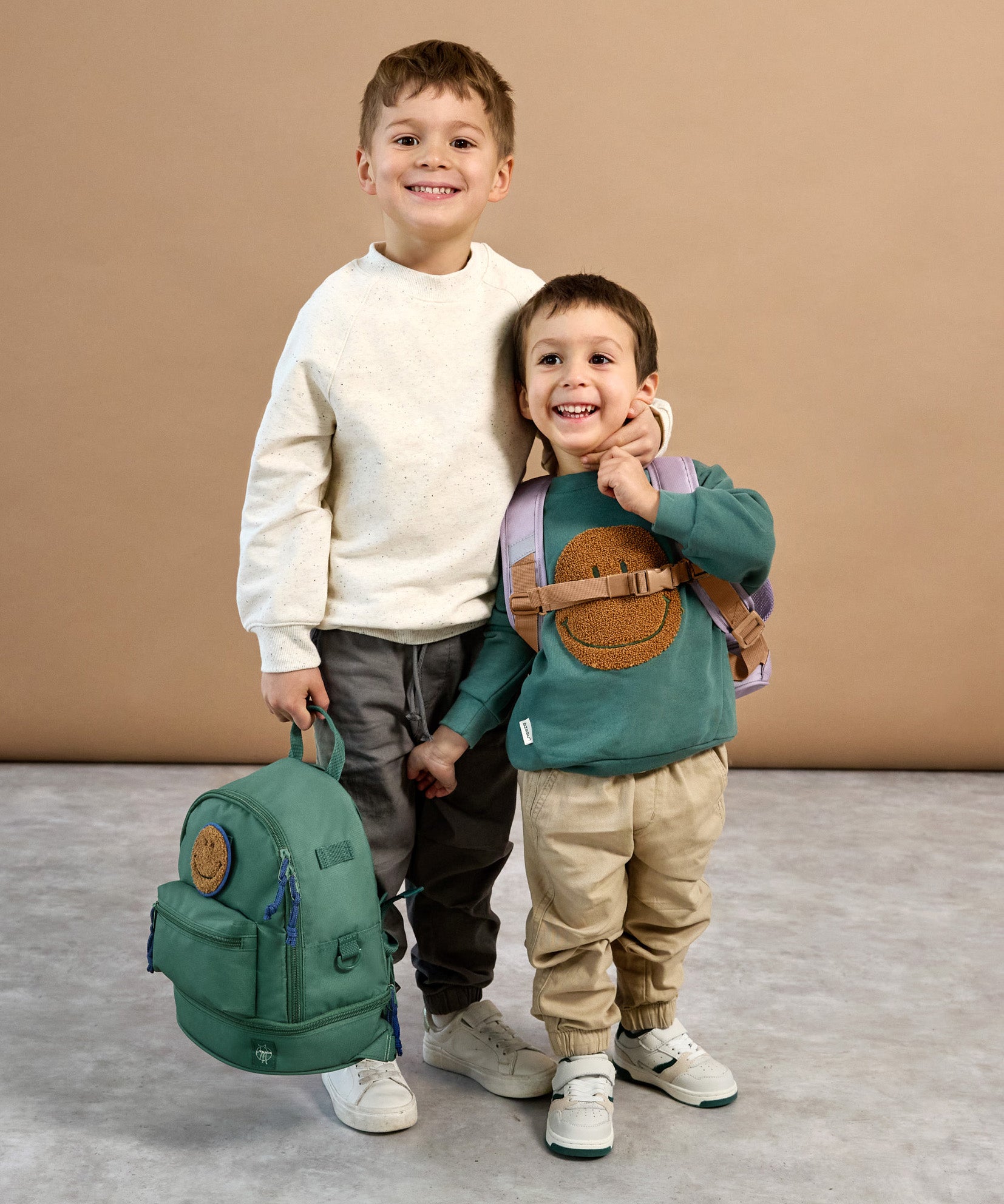 A boy holding the Lassig mini backpack for kids in a dark green colour, with his arm around a younger boy who's wearing the lilac version.