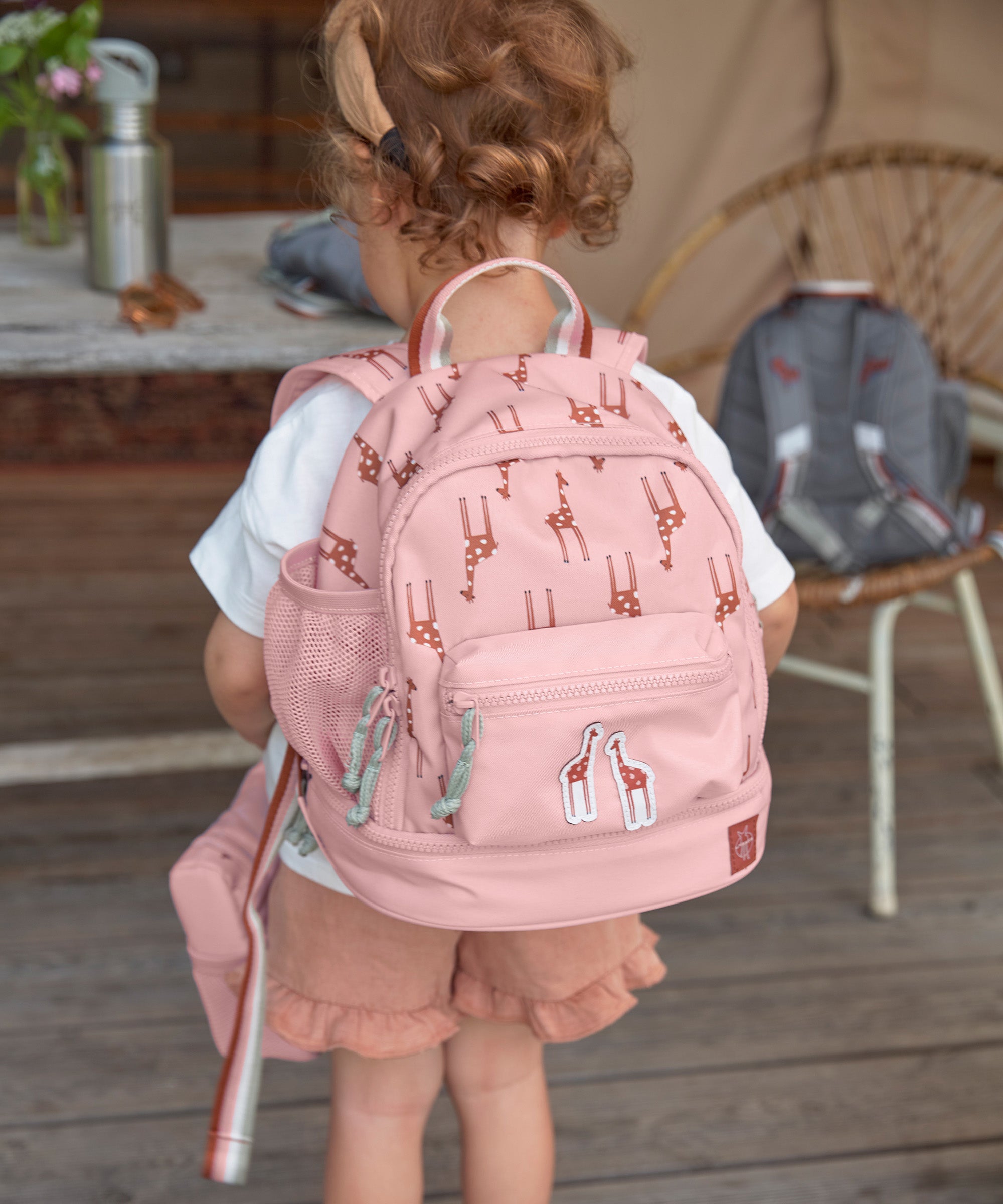 A little girl wearing the Lassig kids mini backpack in a Safari Giraffe pink print.