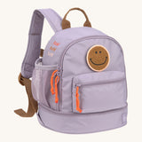 The Lassig mini kids backpack in a lilac colour with orange zips and a smiley face patch.