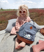 A little girl in sunglasses holding her Lassig kids mini backpack with a tractor on the front.