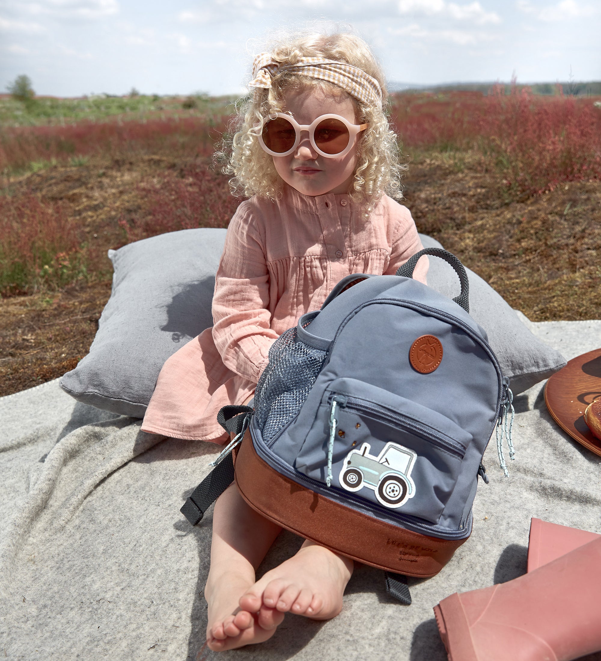 A little girl in sunglasses holding her Lassig kids mini backpack with a tractor on the front.