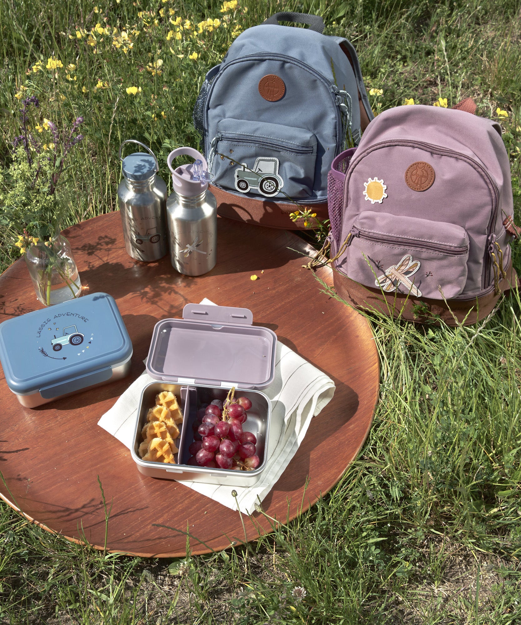 Two Lassig mini kids backpacks, packed ready for a picnic on the grass.