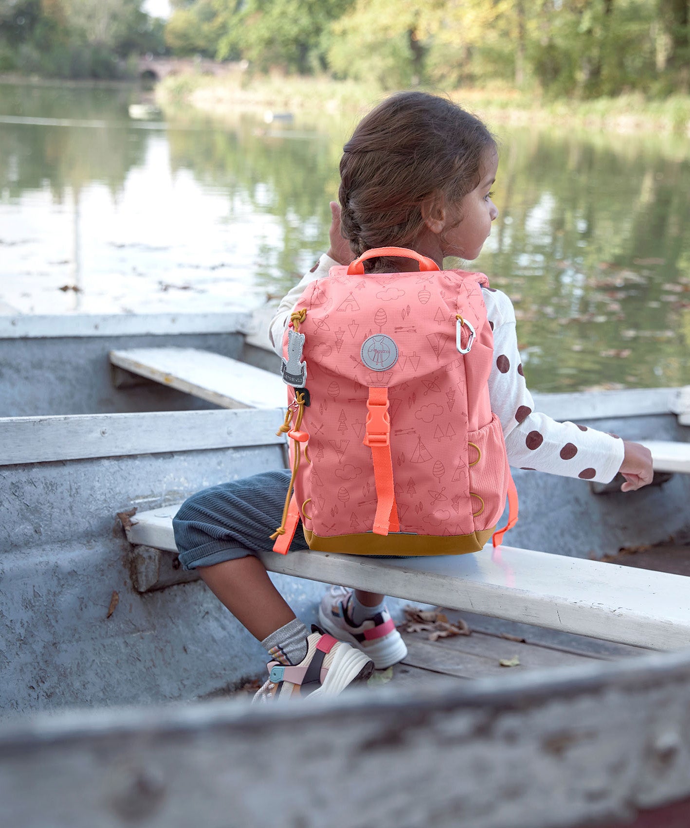 A child on a boat near a river and wearing the Lassig kids mini adventure backpack in rose.