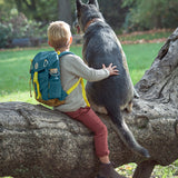 A little boy sitting on a tree branch with a big dog, wearing the Lassig kids mini outdoor backpack in Adventure Blue.