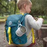 A little boy wearing the Lassig kids mini outdoor backpack in Adventure Blue, with a camping mug attached to it.