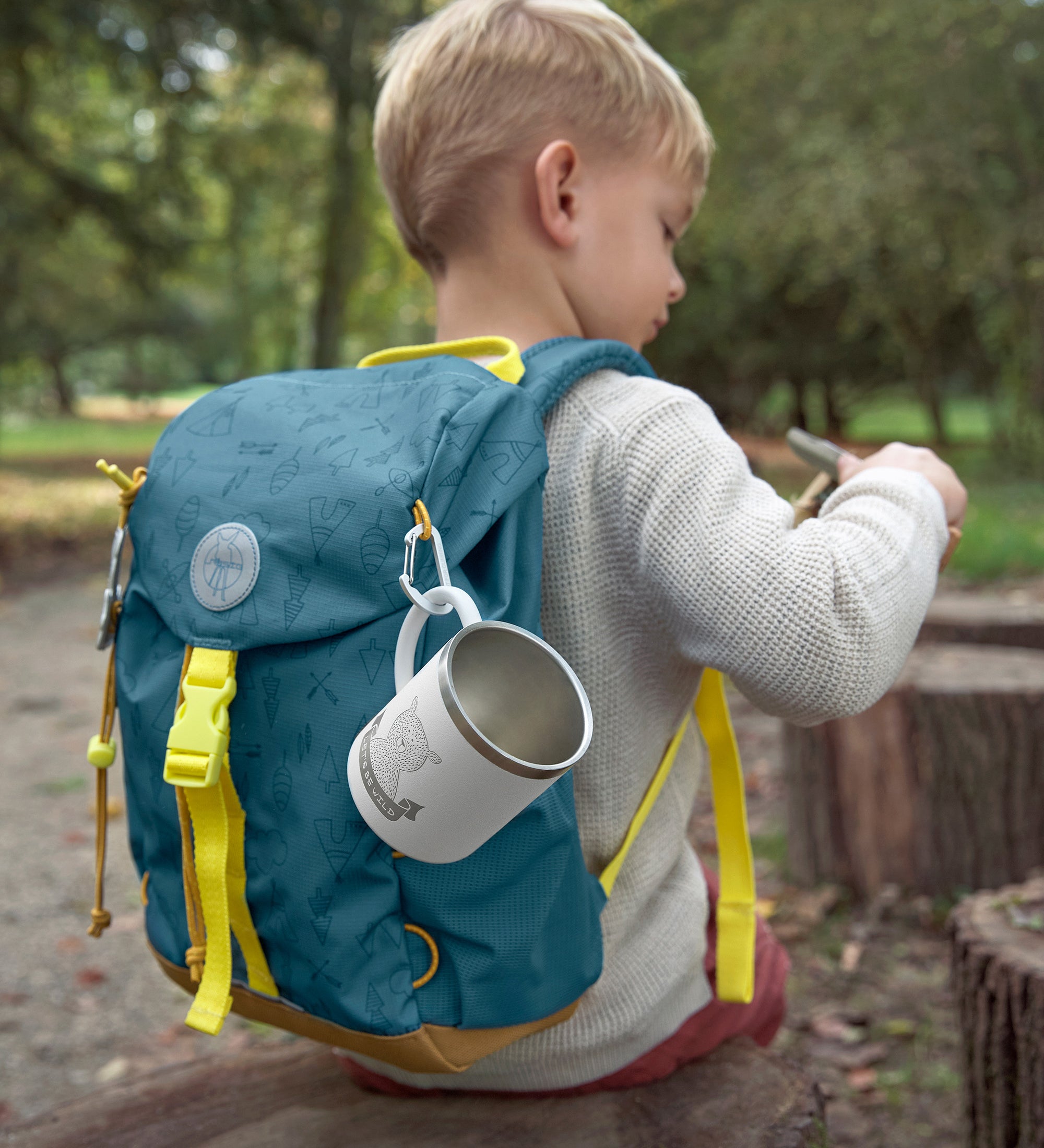 A little boy wearing the Lassig kids mini outdoor backpack in Adventure Blue, with a camping mug attached to it.