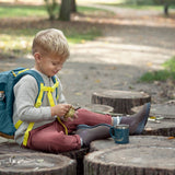A little boy sat on some logs, wearing the Lassig kids mini outdoor backpack in Adventure Blue.