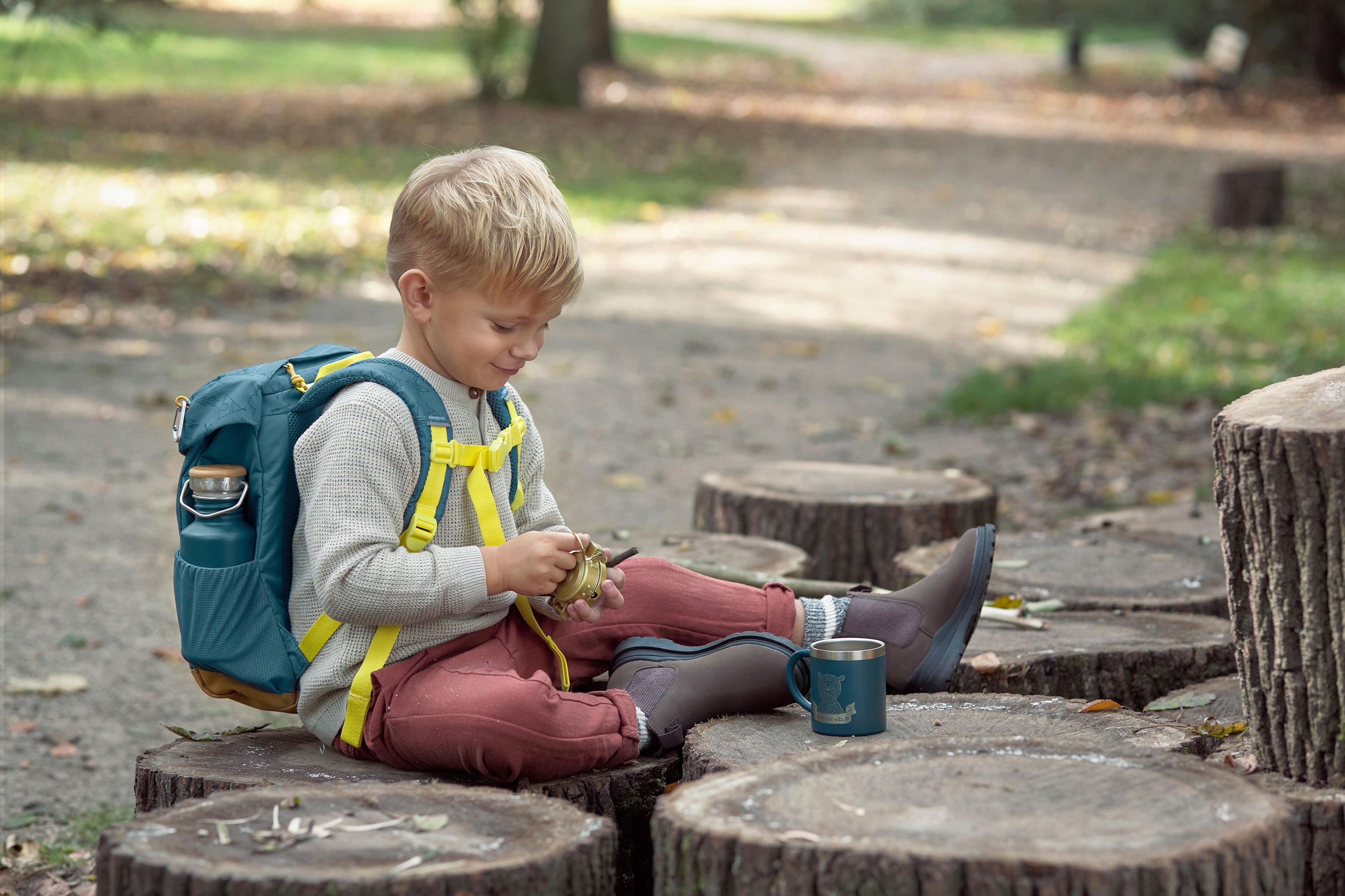 A little boy sat on some logs, wearing the Lassig kids mini outdoor backpack in Adventure Blue.