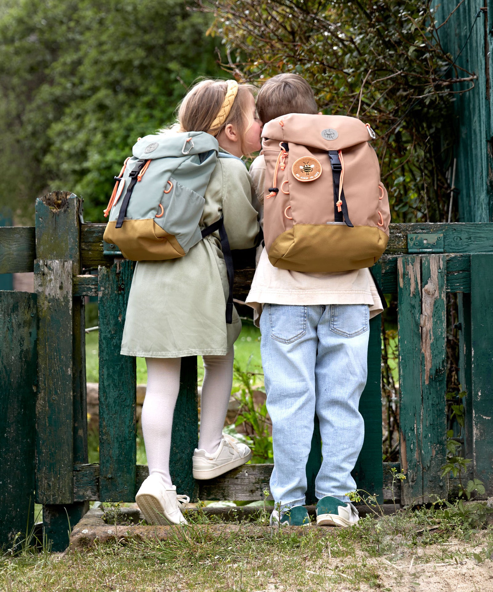 Two kids wearing their Lassig kids mini outdoor backpacks, leaning against a gate.
