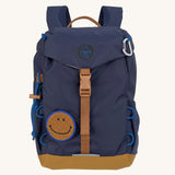The Lassig kids mini outdoor backpack in navy blue, with a brown strap and buckle, and a smiley face patch.