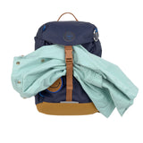 The Lassig kids mini outdoor backpack in navy blue, with a coat secured under the brown strap.