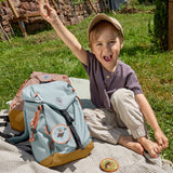 A little boy sat in the grass next to his Lassig kids mini outdoor backpack in blue, and holding one of the customisable bag patches.