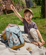 A little boy sat in the grass next to his Lassig kids mini outdoor backpack in blue, and holding one of the customisable bag patches.