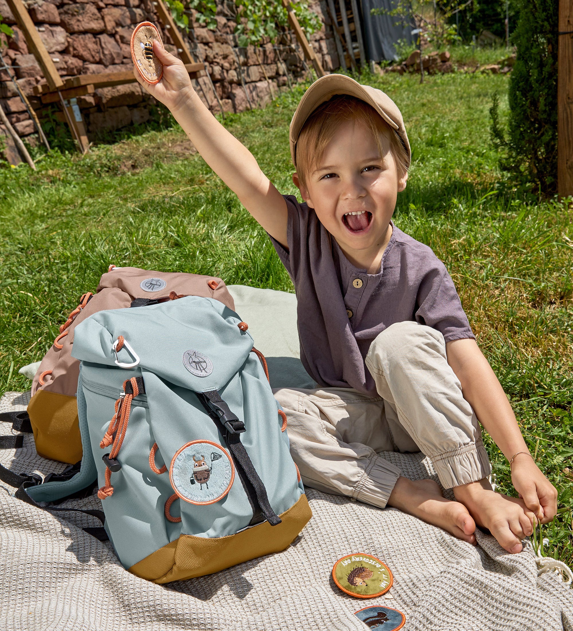 A little boy sat in the grass next to his Lassig kids mini outdoor backpack in blue, and holding one of the customisable bag patches.