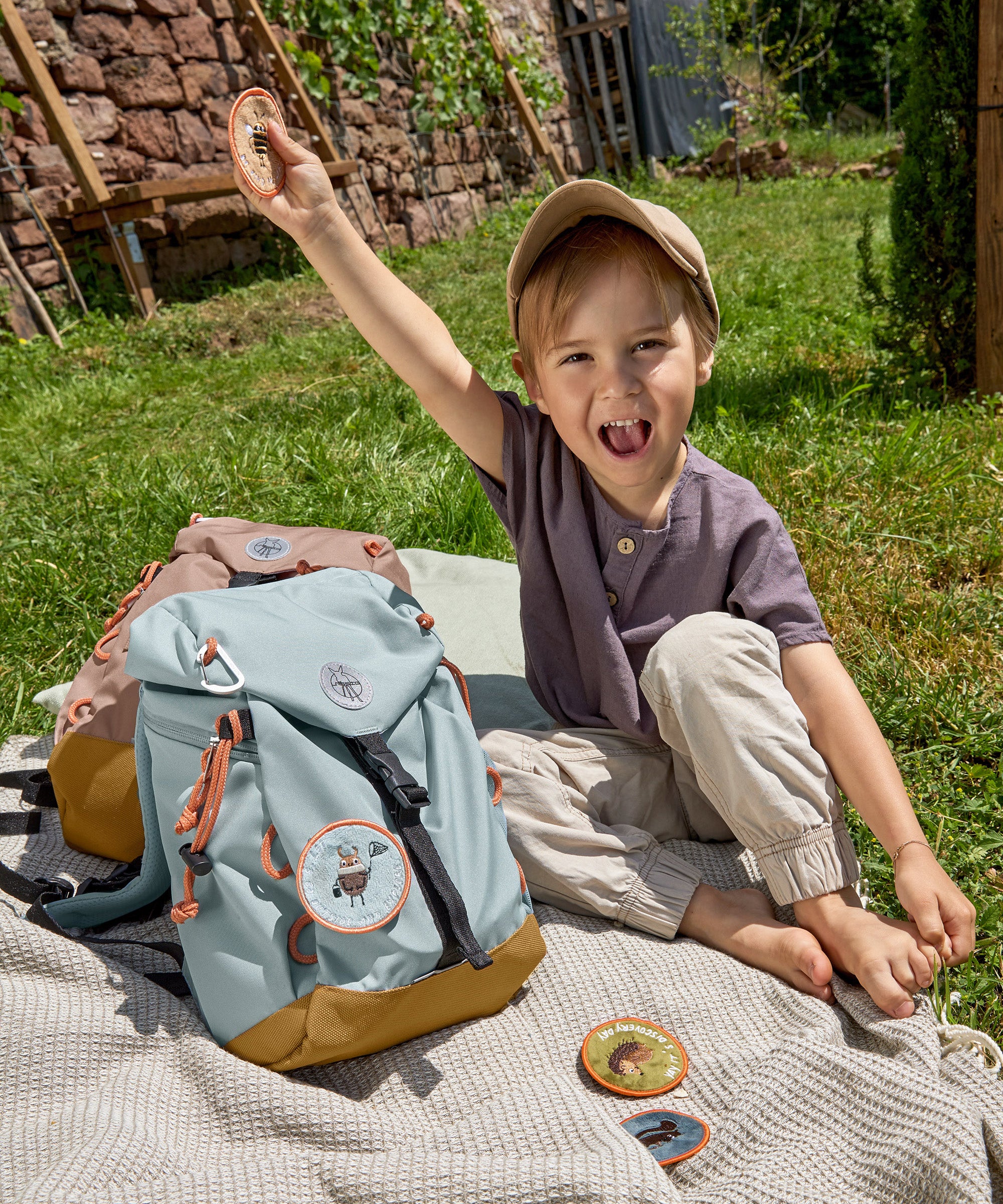 A little boy sat in the grass next to his Lassig kids mini outdoor backpack in blue, and holding one of the customisable bag patches.