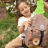 A little girl pulling out the protective waterproof cover from her Lassig mini outdoor backpack in hazelnut.