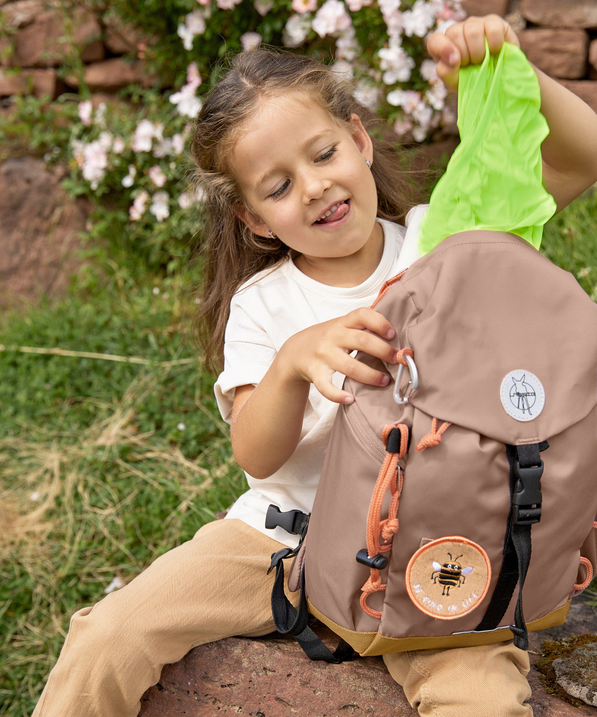A little girl pulling out the protective waterproof cover from her Lassig mini outdoor backpack in hazelnut.
