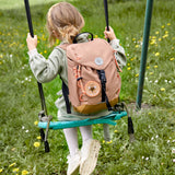 A child on a swing, wearing the Lassig kids mini outdoor backpack in a hazelnut light brown colour