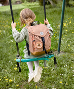 A child on a swing, wearing the Lassig kids mini outdoor backpack in a hazelnut light brown colour