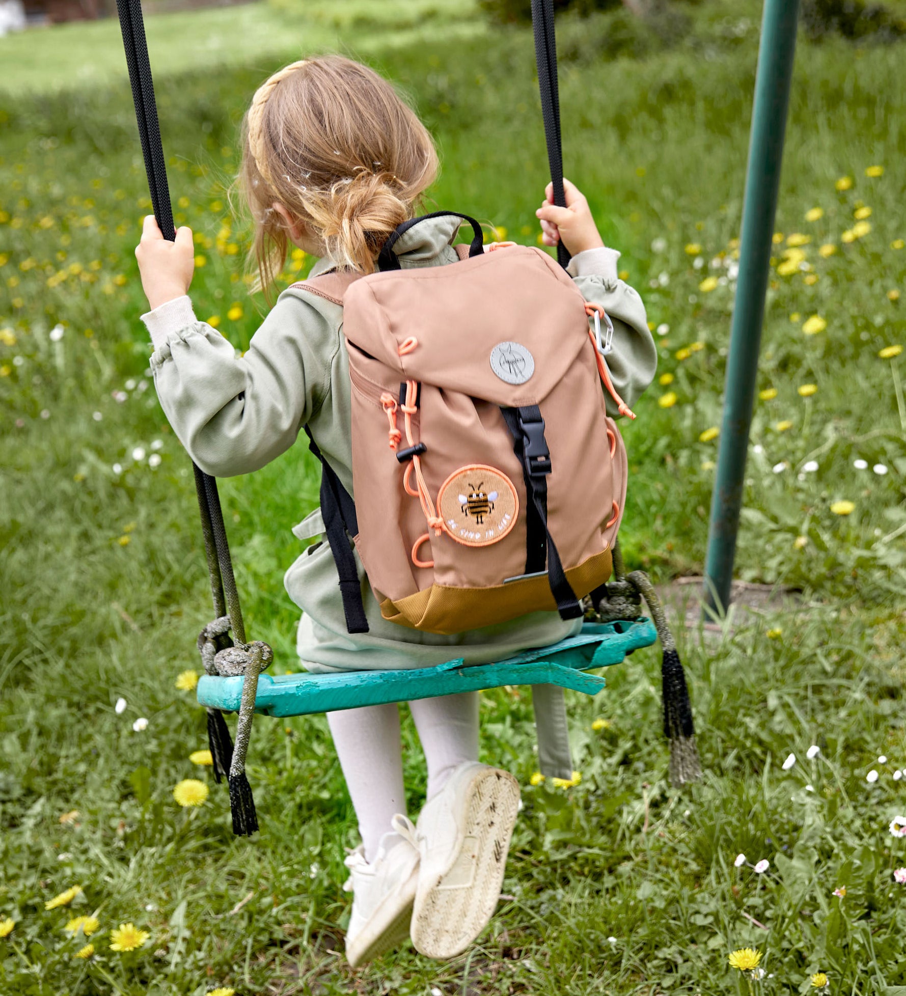 A child on a swing, wearing the Lassig kids mini outdoor backpack in a hazelnut light brown colour