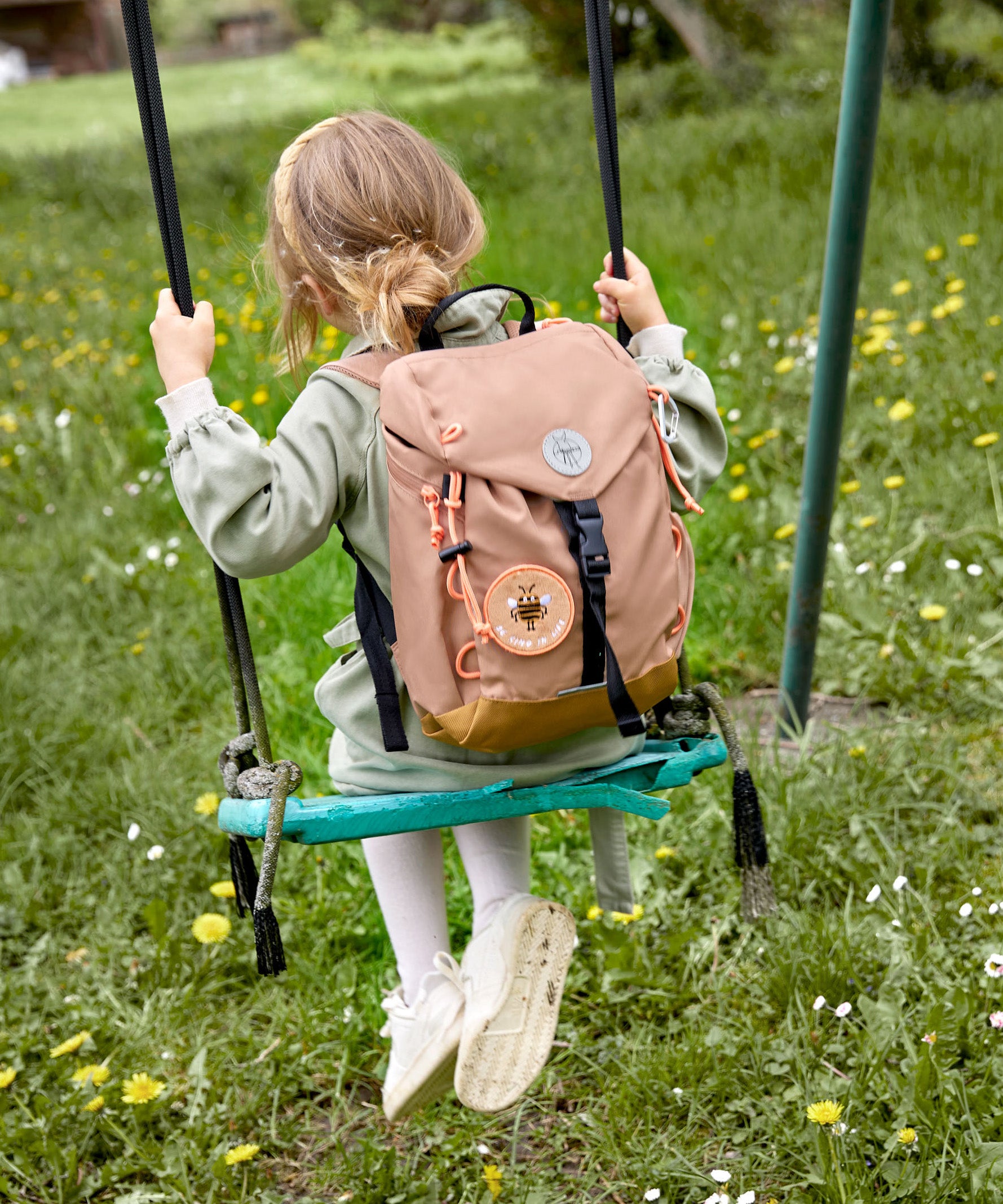 A child on a swing, wearing the Lassig kids mini outdoor backpack in a hazelnut light brown colour