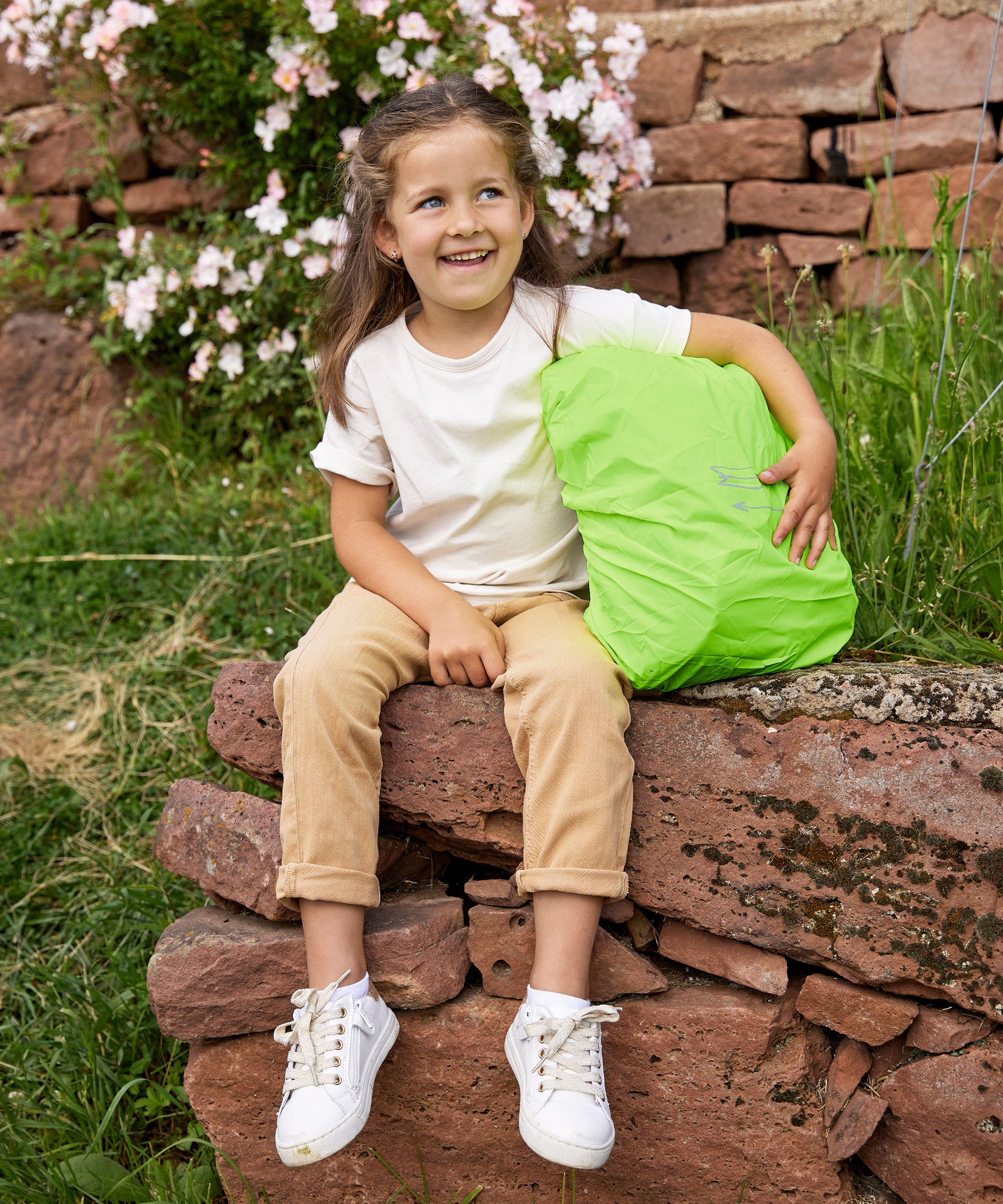 A girl sitting on a wall with her Lassig kids mini outdoor backpack, with the waterproof cover on it.
