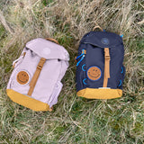 Two Lassig kids mini outdoor backpacks, one navy and one lilac, on a field.