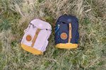 Two Lassig kids mini outdoor backpacks, one navy and one lilac, on a field.