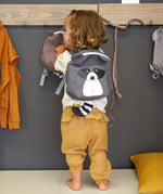 A child wearing the Lassig tiny kids backpack in grey, with a racoon face printed on and 3D ears and tail.