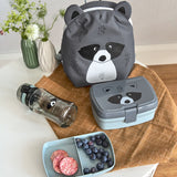 The Lassig tiny kids backpack in grey, with a racoon face printed on and 3D ears and tail, on a table with a lunchbox and a water bottle.