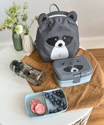The Lassig tiny kids backpack in grey, with a racoon face printed on and 3D ears and tail, on a table with a lunchbox and a water bottle.