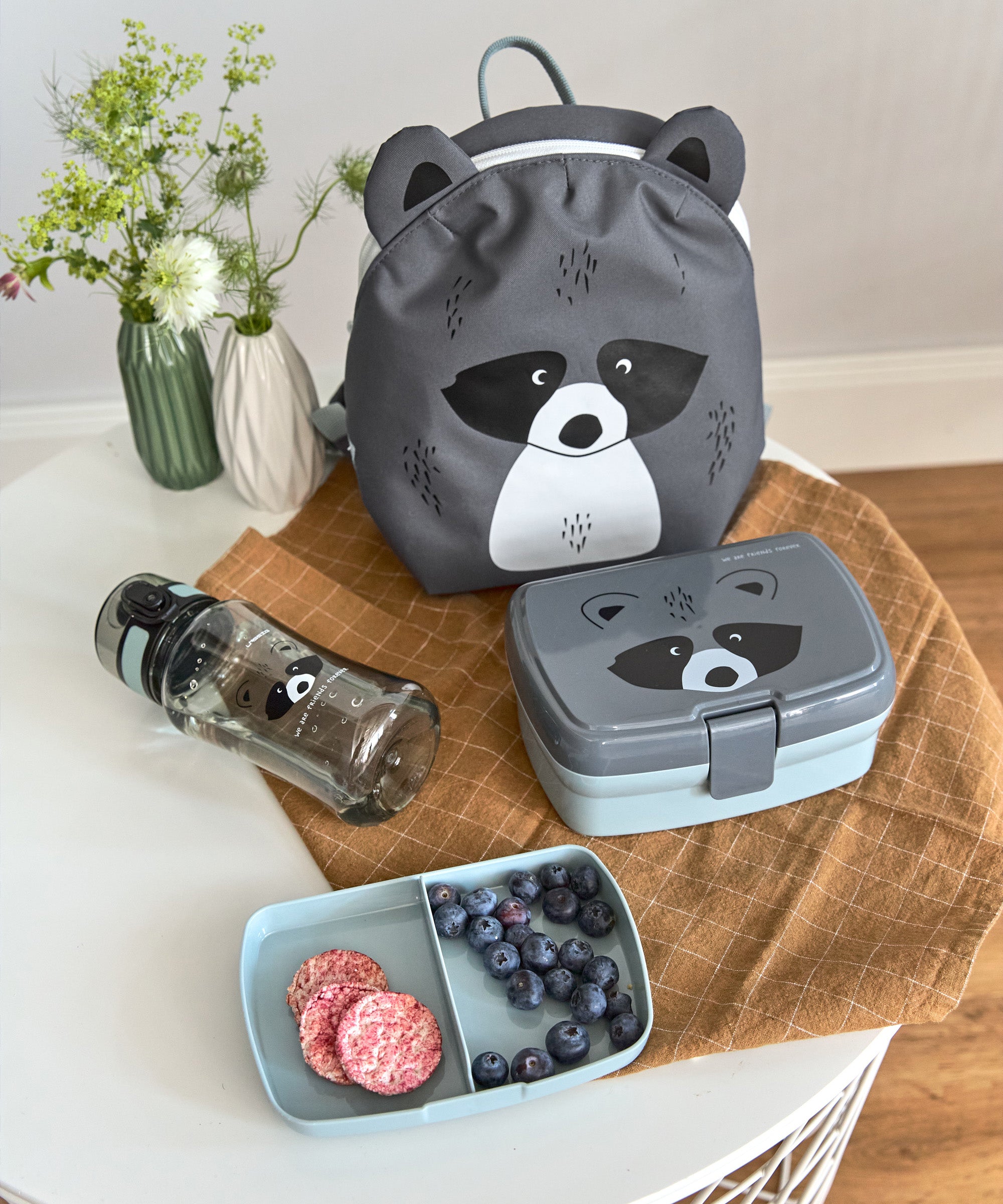The Lassig tiny kids backpack in grey, with a racoon face printed on and 3D ears and tail, on a table with a lunchbox and a water bottle.