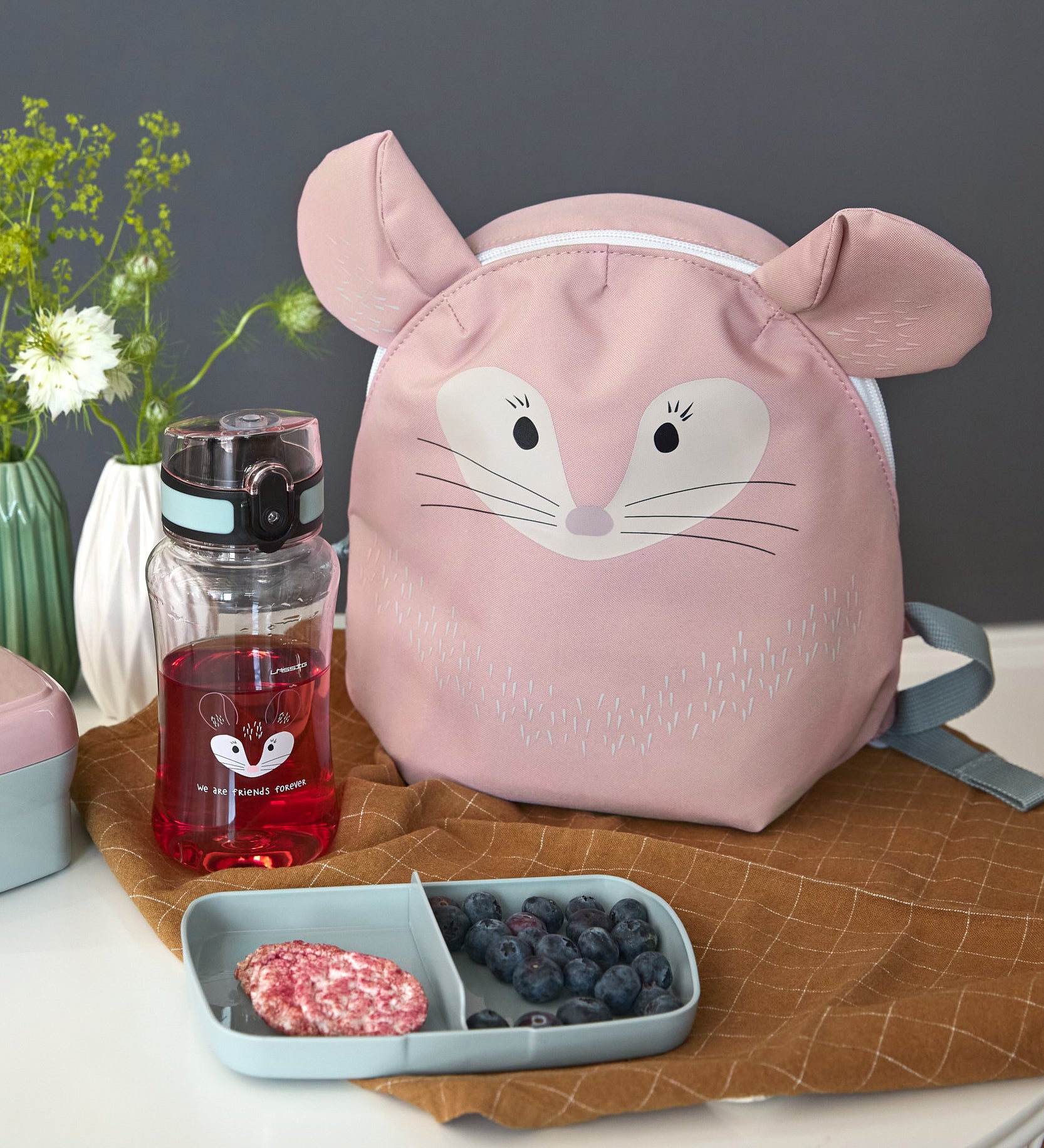 The Lassig 'About Friends' tiny backpack in light pink, with a chinchilla face and 3D ears, on a table next to a lunch box and water bottle.