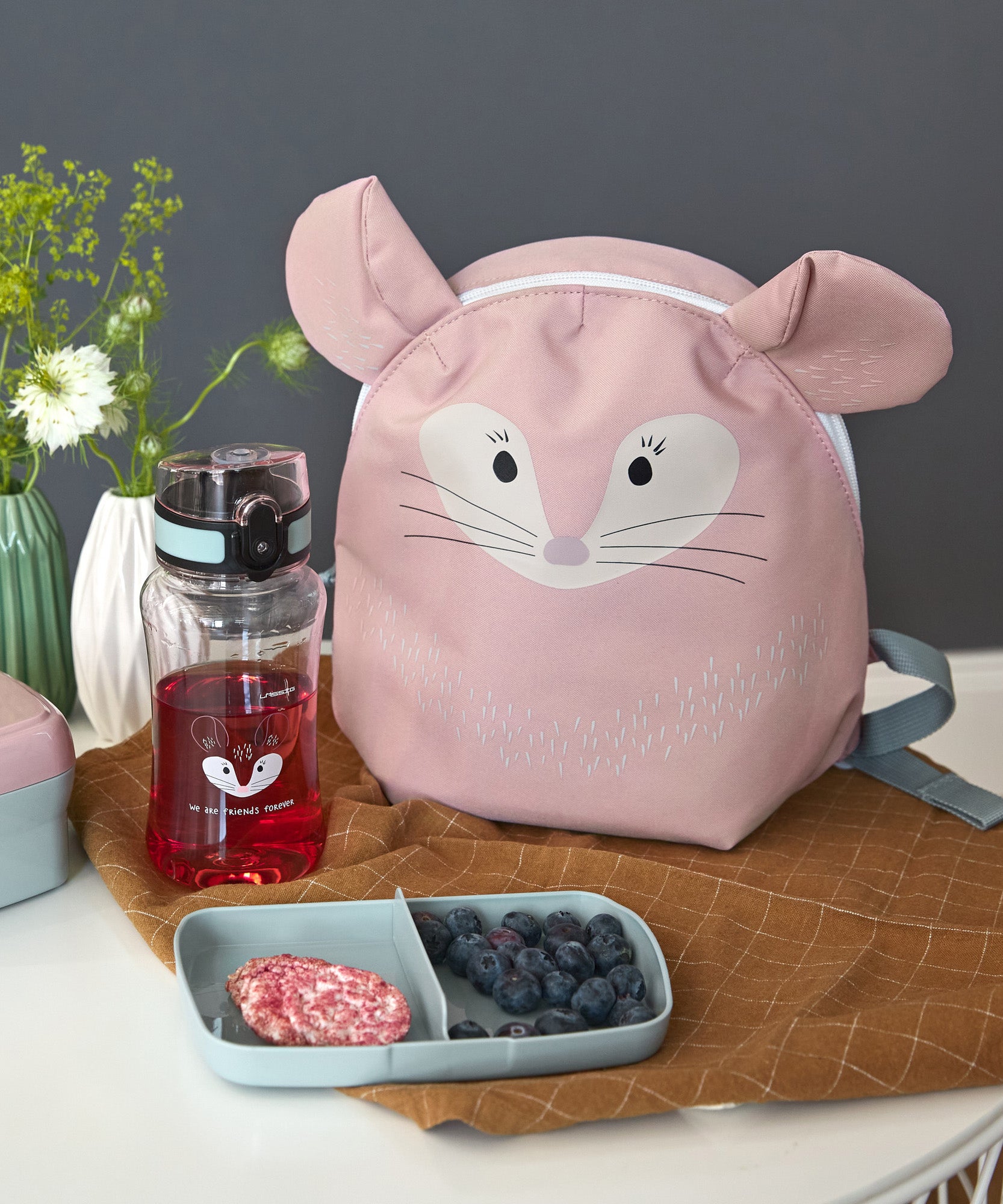 The Lassig 'About Friends' tiny backpack in light pink, with a chinchilla face and 3D ears, on a table next to a lunch box and water bottle.