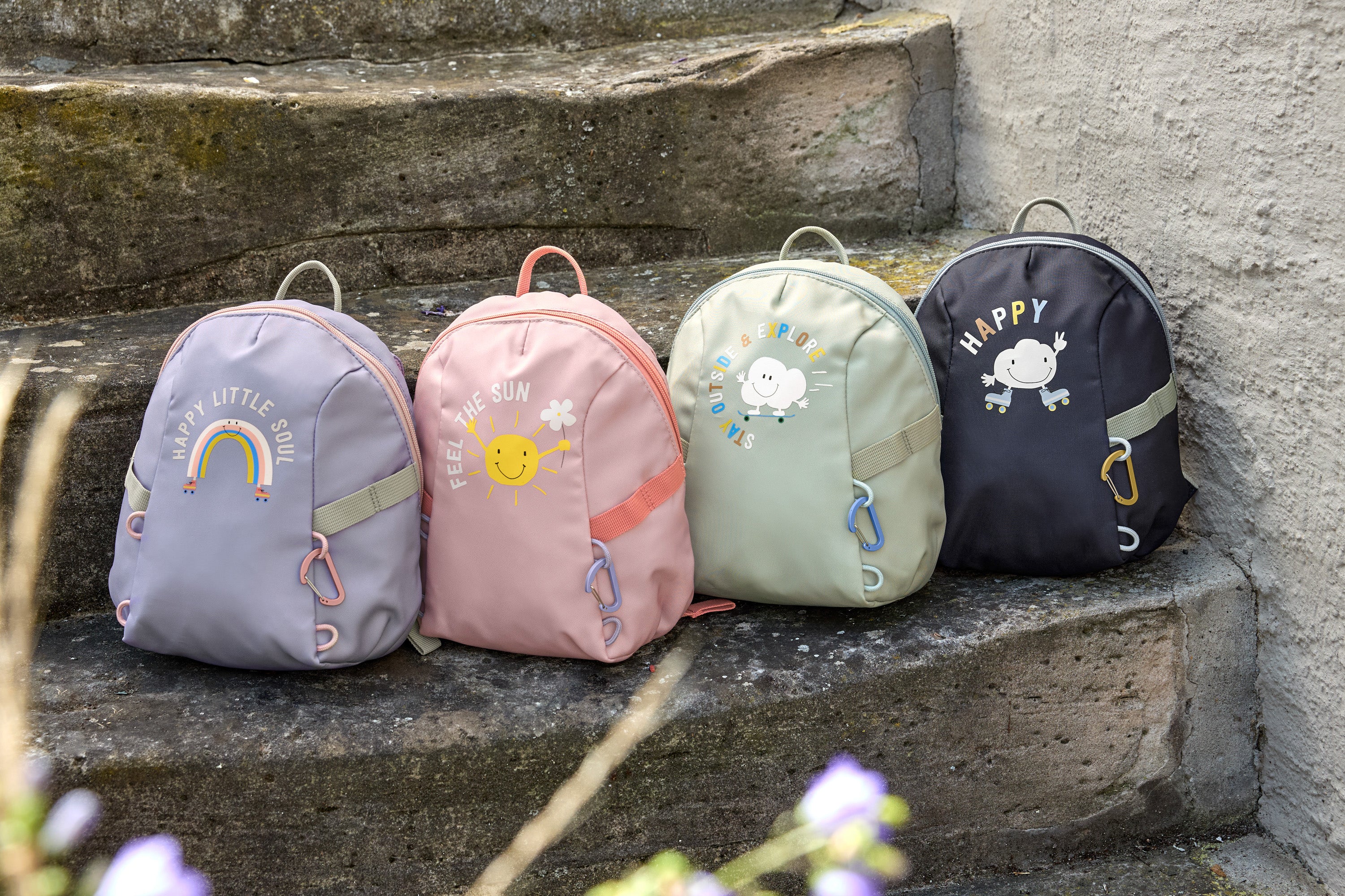 Four Lassig kids tiny backpacks placed on stairs, one is in a light green khaki, one is navy blue, one is pink, and one is pastel purple.