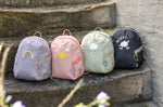 Four Lassig kids tiny backpacks placed on stairs, one is in a light green khaki, one is navy blue, one is pink, and one is pastel purple.