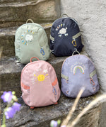 Four Lassig kids tiny backpacks placed on stairs, one is in a light green khaki, one is navy blue, one is pink, and one is pastel purple.