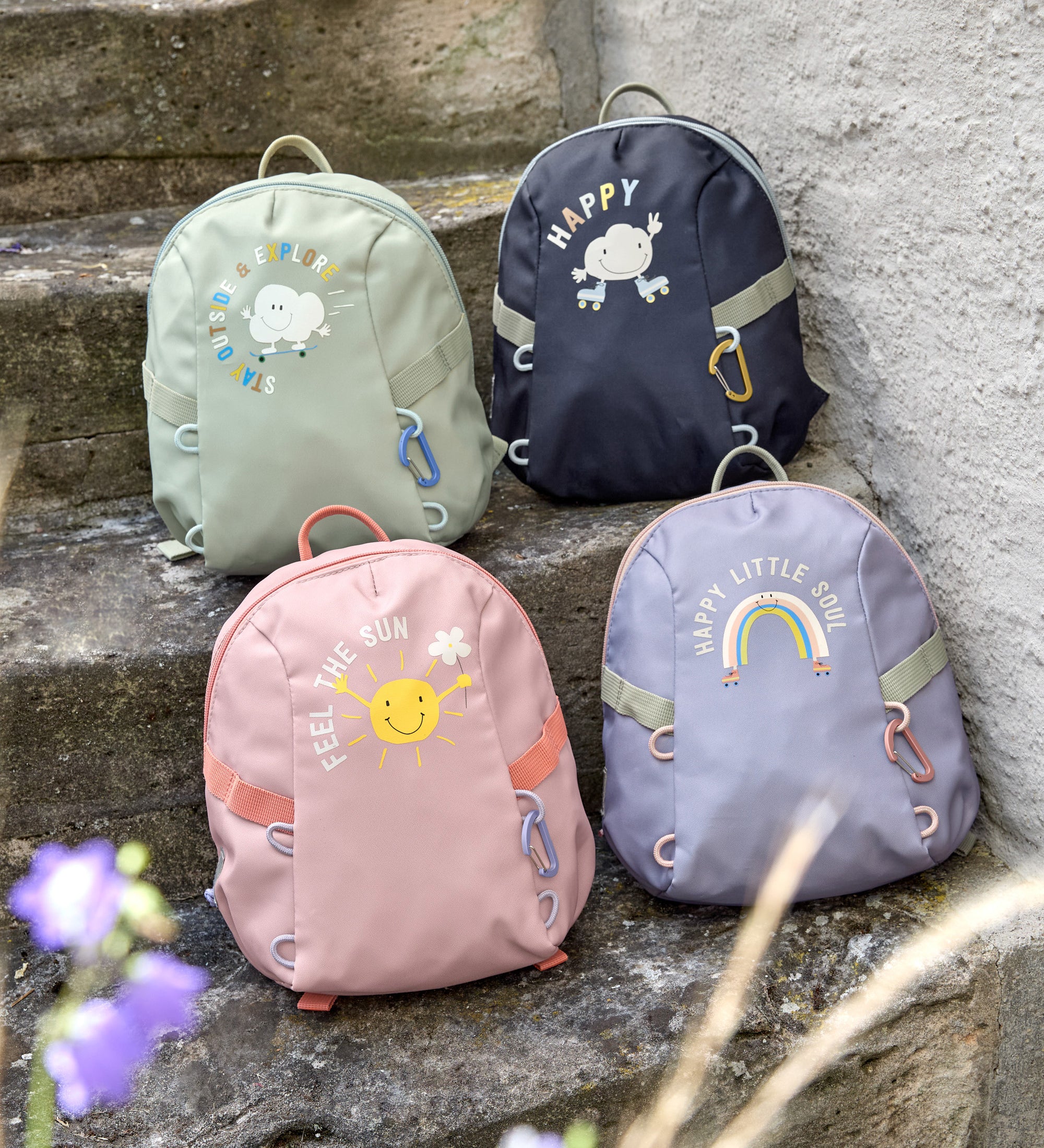 Four Lassig kids tiny backpacks placed on stairs, one is in a light green khaki, one is navy blue, one is pink, and one is pastel purple.