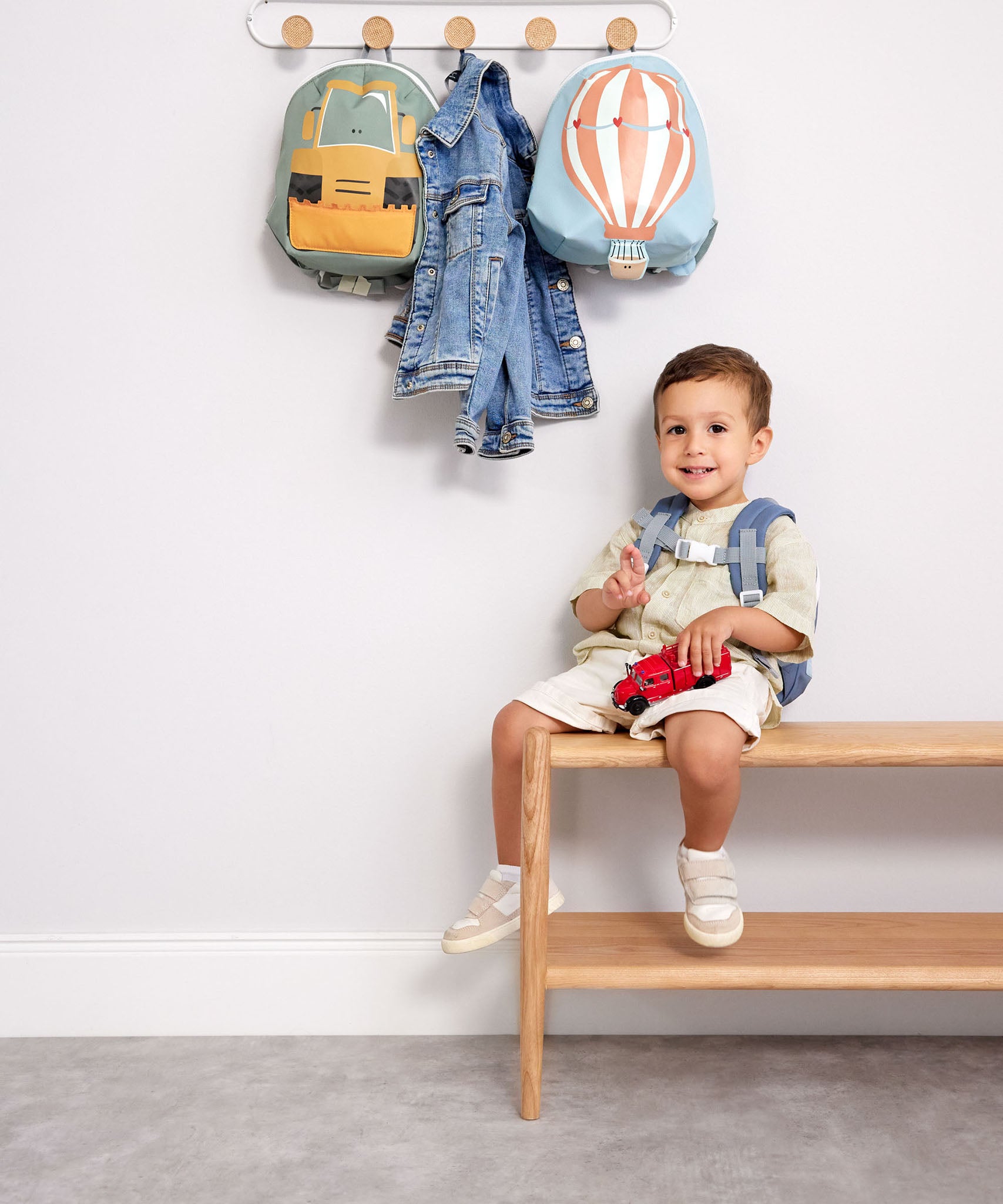 A child sitting on a wooden bench with a digger and balloon character Lassig tiny backpacks hanging from hooks above.