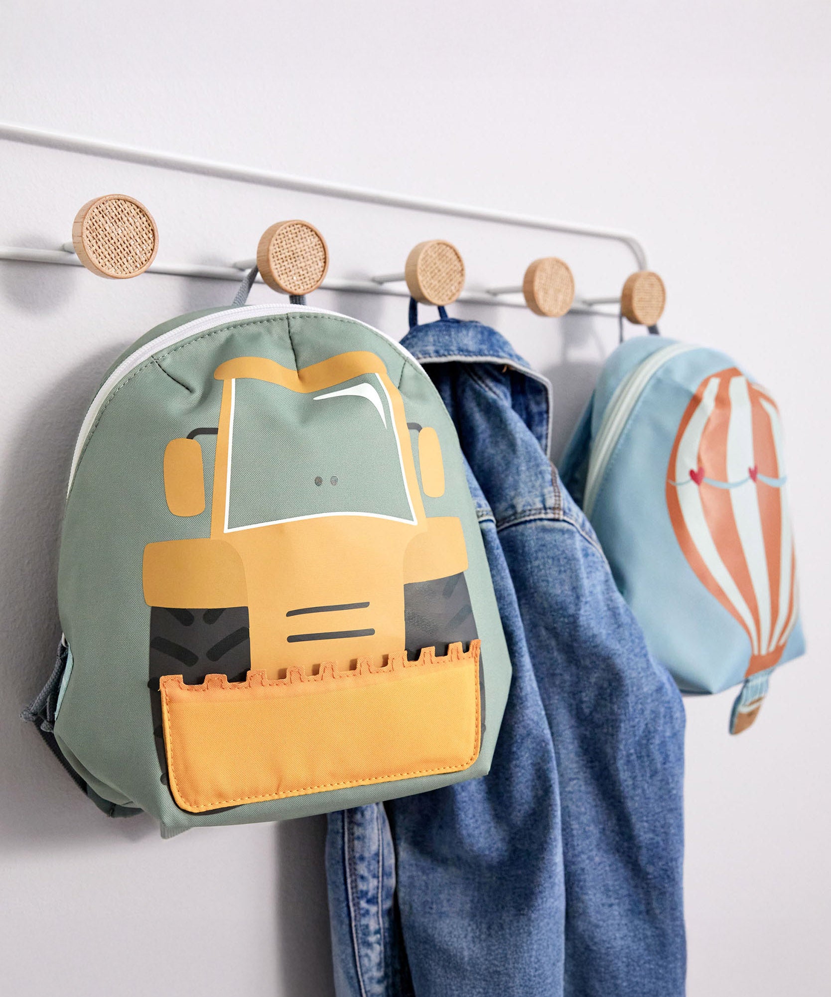 Lassig kids tiny backpack in digger and balloon design hanging from hooks with a denim jacket inbetween