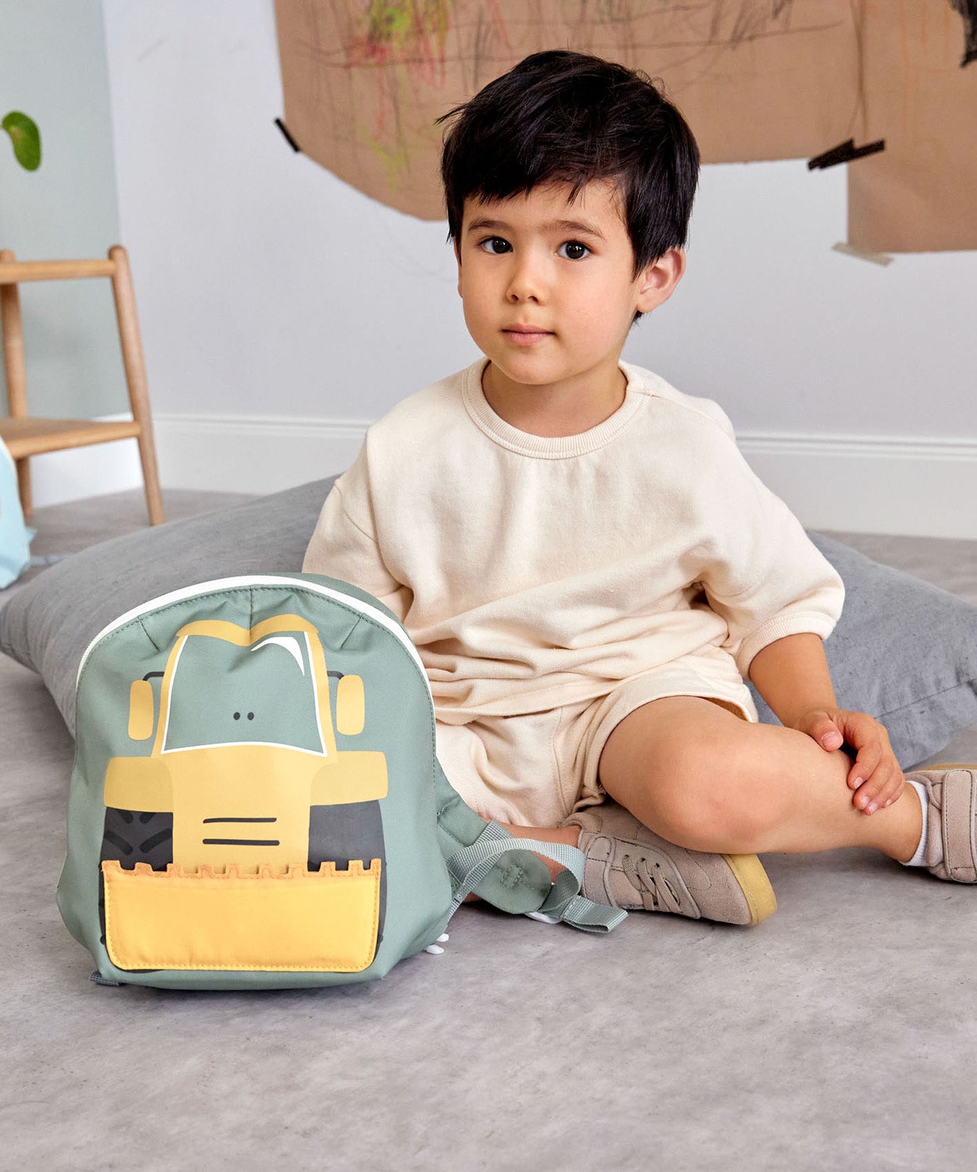A child sitting on a carpet floor in a playroom next to the Lassig kids tiny digger backpack