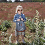A girl in a sunny field, holding her Lassig mini backpack in blue with a tractor on it.