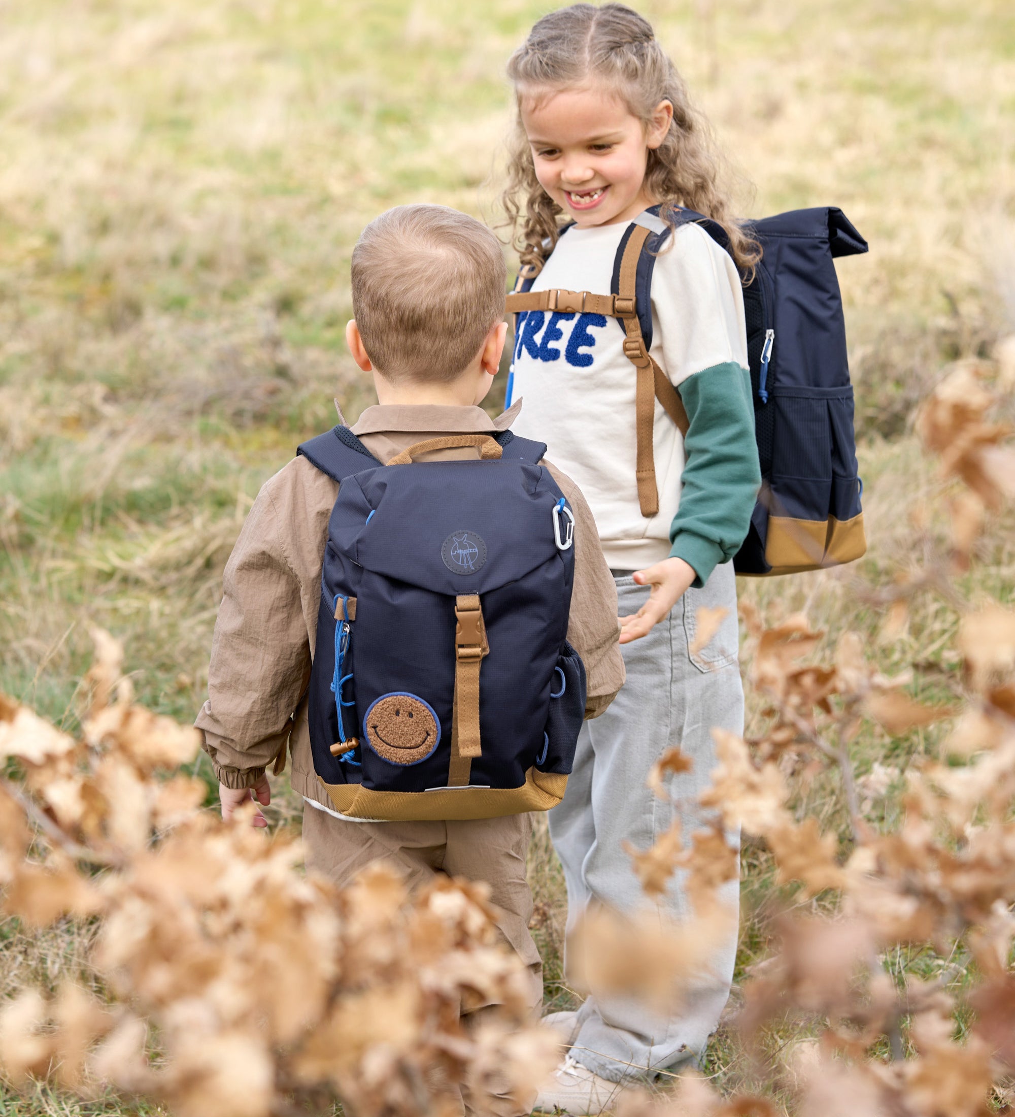 Two children wearing Lassig mini backpacks in navy blue, in a green field.
