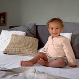 A baby sat on a bed, wearing the Lassig organic cotton muslin long sleeved top for babies and toddlers, in a beautiful pale powder pink colour.