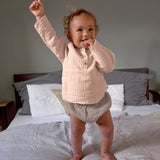 A baby standing on a bed, wearing the Lassig organic cotton muslin long sleeved shirt for babies and toddlers in a beautiful pale powder pink colour