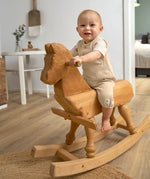 A little toddler sat on a wooden rocking horse, wearing the Lassig organic cotton muslin jumpsuit in a natural cream colour.