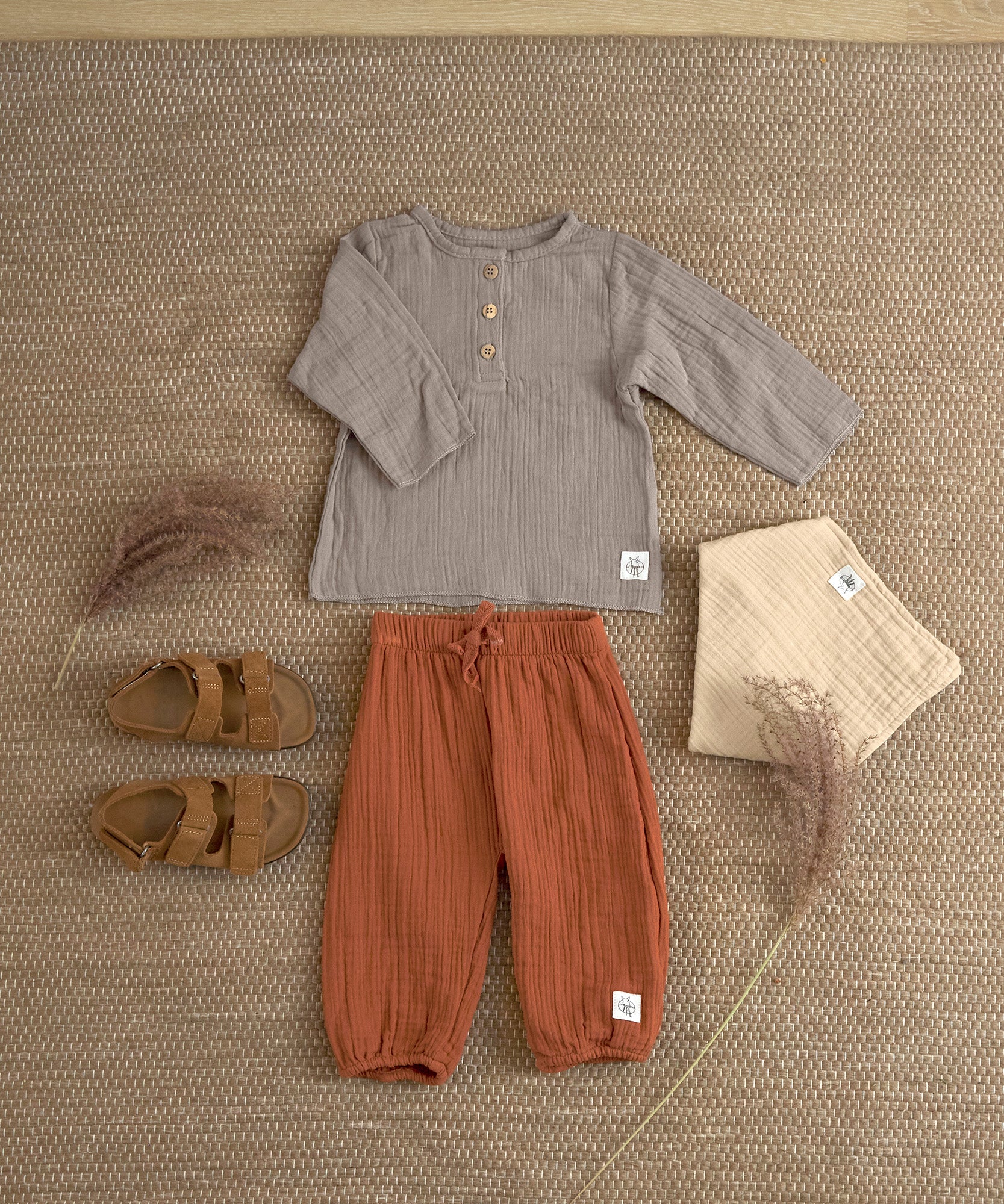 An organic cotton muslin outfit from Lassig, laid out on the floor. It includes the Lassig muslin pants in orange rust.