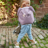 A child standing on a cobbled path and holding the Lassig bunny tiny backpack by the loop handle on the top